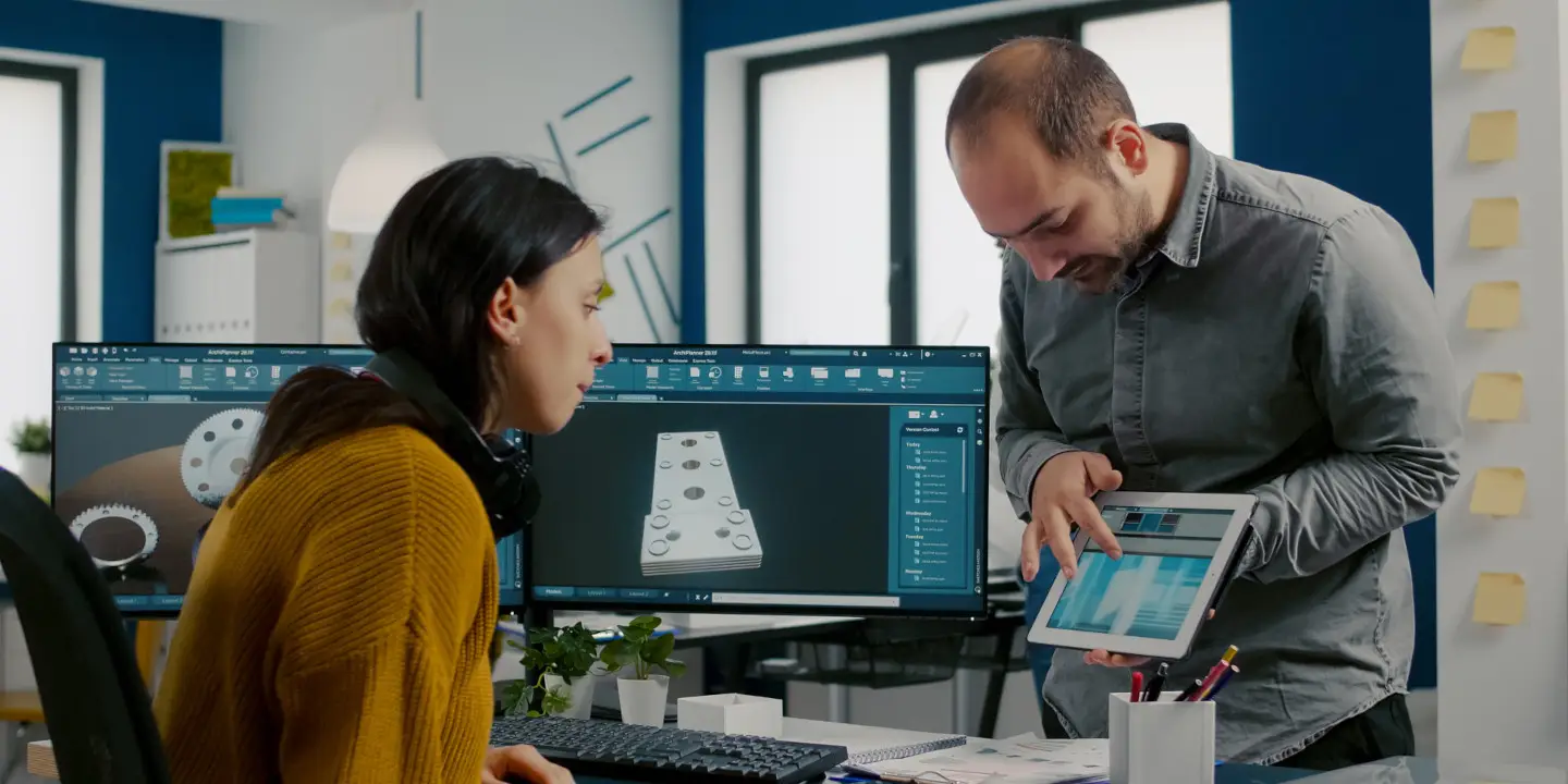 A man showcases a digital blueprint on a tablet, highlighting the Revit design features to a woman seated at a desk adorned with multiple monitors displaying CAD designs in an office environment.