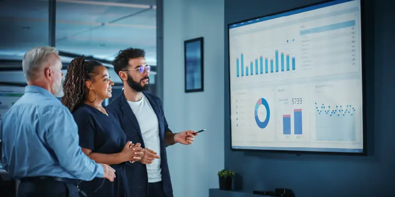 Three people stand in an office, reviewing charts and graphs on a large screen display, analyzing manufacturing sales trends.