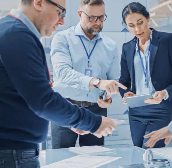 A group of professionals in an office setting discuss documents and data on a tablet laid out on a table.