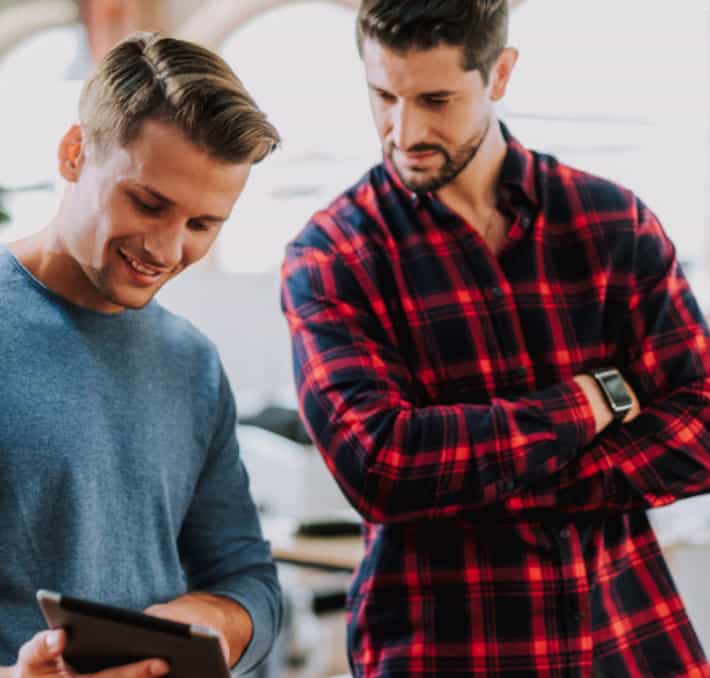 Two men are standing indoors. One, a PLM partner, is holding a tablet and smiling, while the other, wearing a red plaid shirt, looks at the screen with his arms crossed.