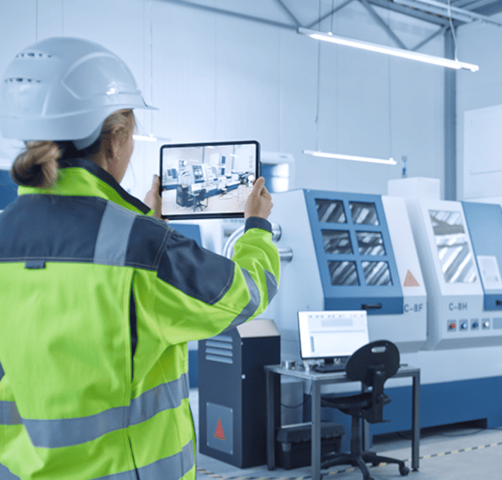 A person in a high-visibility jacket and helmet uses a tablet to capture images in a factory with machinery and monitors in the background.
