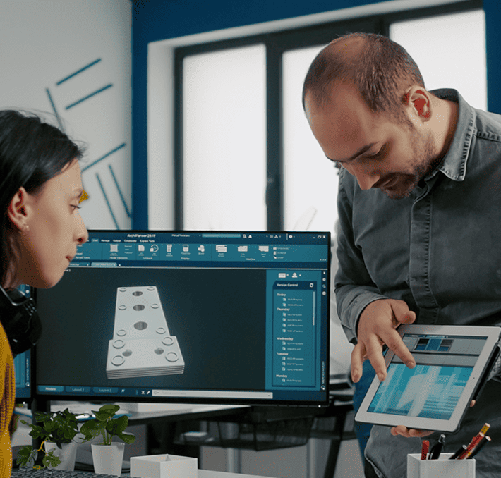 Two people in an office discuss 3D model designs displayed on a computer and tablet. A monitor shows a 3D object.