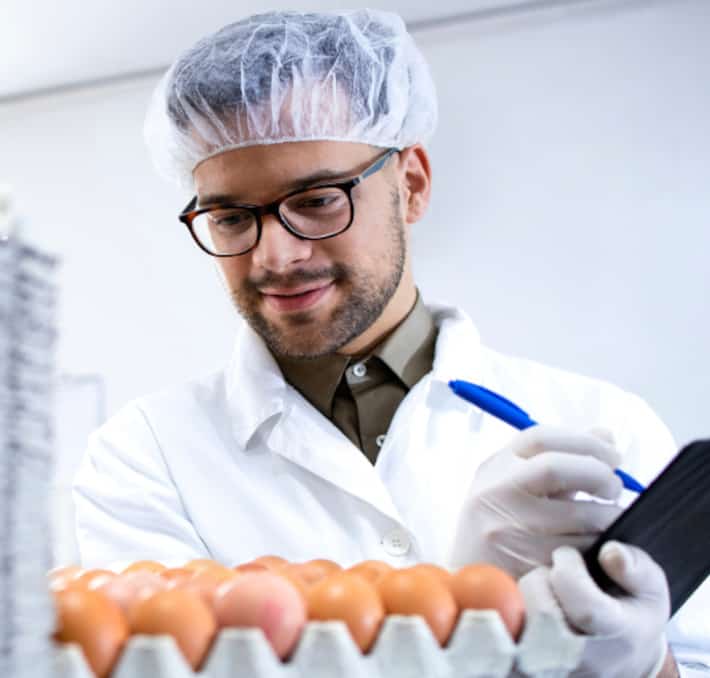 Man in a lab coat and hairnet examines eggs, holding a pen and notepad, much like a chef perfecting his almondy creations.