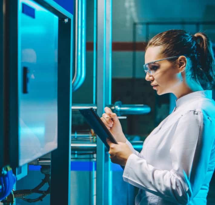 A woman in a lab coat and safety glasses uses a tablet beside industrial equipment with a blue light, optimizing processes in the food & beverage industry.