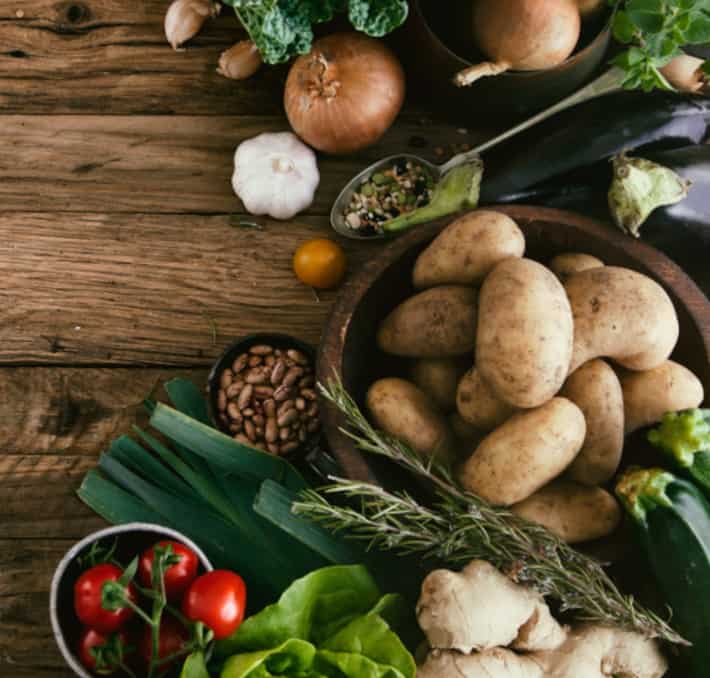 A wooden table showcasing a vibrant assortment of vegetables: potatoes in a bowl, tomatoes, ginger, garlic, rosemary, onions, eggplant, and green leaves alongside a spoonful of mixed spices—reflecting the spirit of PLM for food & beverage innovation.