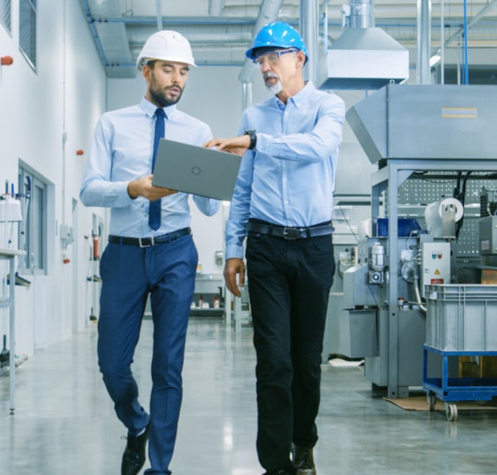 Two men in hard hats and shirts walk through an industrial facility discussing a laptop. One wears a white hard hat, the other a blue one. Machinery is visible in the background.