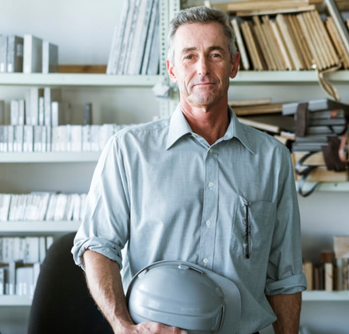 A man in a gray shirt holds a gray hard hat, standing in an office with shelves filled with an extensive medical equipment catalog alongside books and files.