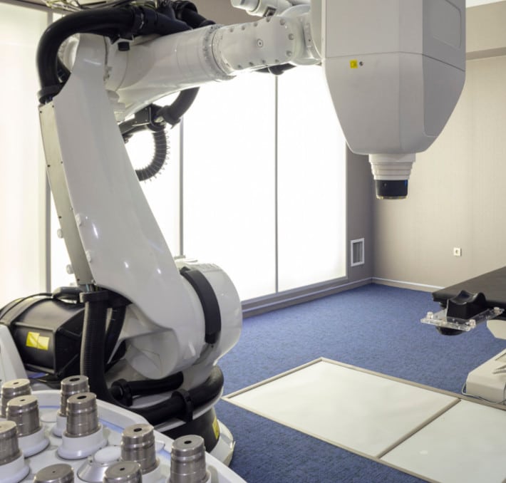 In a bright room, a robotic medical device with an articulated arm and large overhead component showcases advanced medical equipment. Multiple cylindrical parts are neatly arranged on a table in the foreground, highlighting its efficiency and minimizing total cost of ownership (TCO).