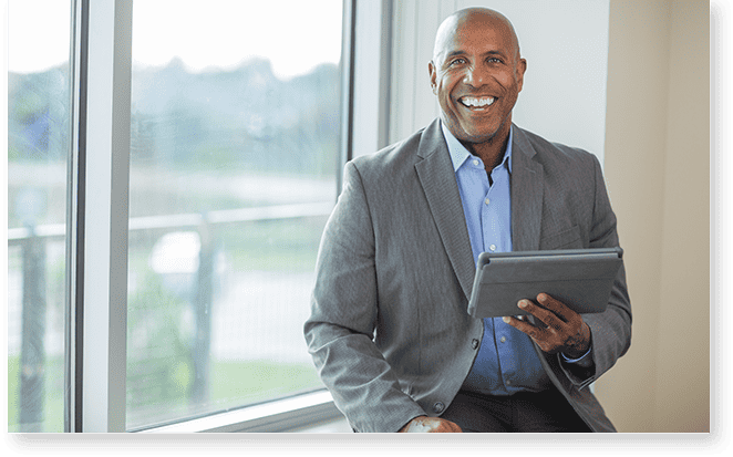 A smiling man in a suit sits by a window, holding a tablet with revalize support, as the blurred outdoor scenery adds to his focused demeanor.