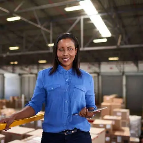 A person in a blue shirt holds a tablet and stands in a warehouse filled with stacked boxes, efficiently managing inventory using advanced manufacturing software.