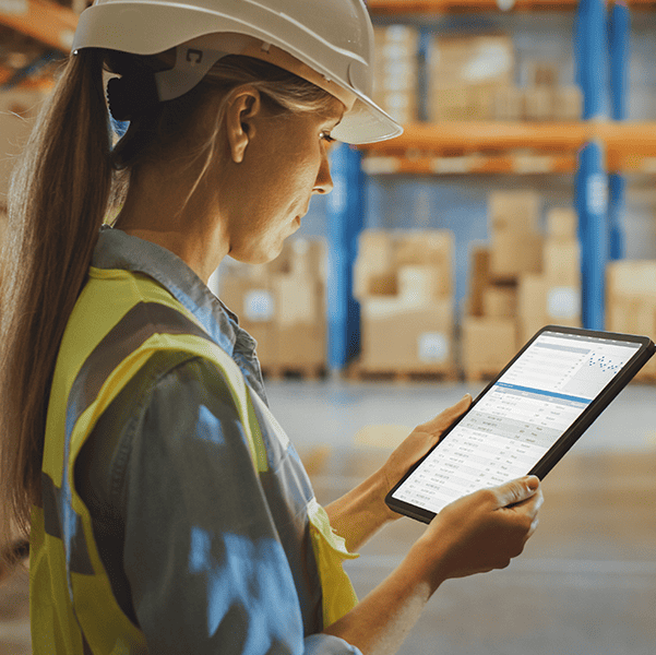 A worker in a hard hat and safety vest efficiently manages inventory on a tablet in a warehouse stacked with boxes, utilizing eRep for Distributors to streamline operations.