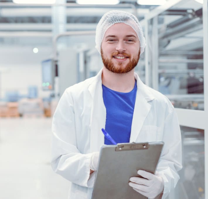 A person in a white lab coat and hairnet stands with a clipboard in an industrial lab, meticulously reviewing the latest research on alternative proteins.
