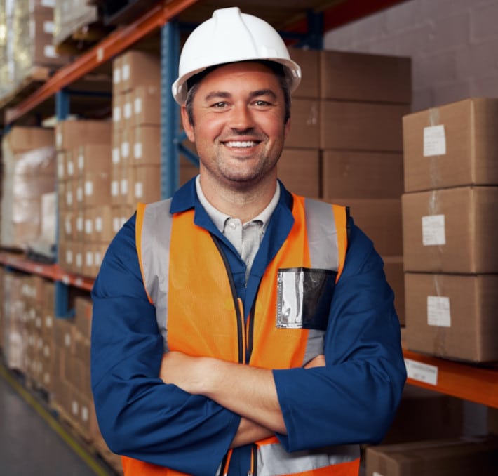 A man in a white hard hat and orange safety vest stands with arms crossed in a warehouse filled with stacked cardboard boxes, overseeing the efficient package design with PLM.