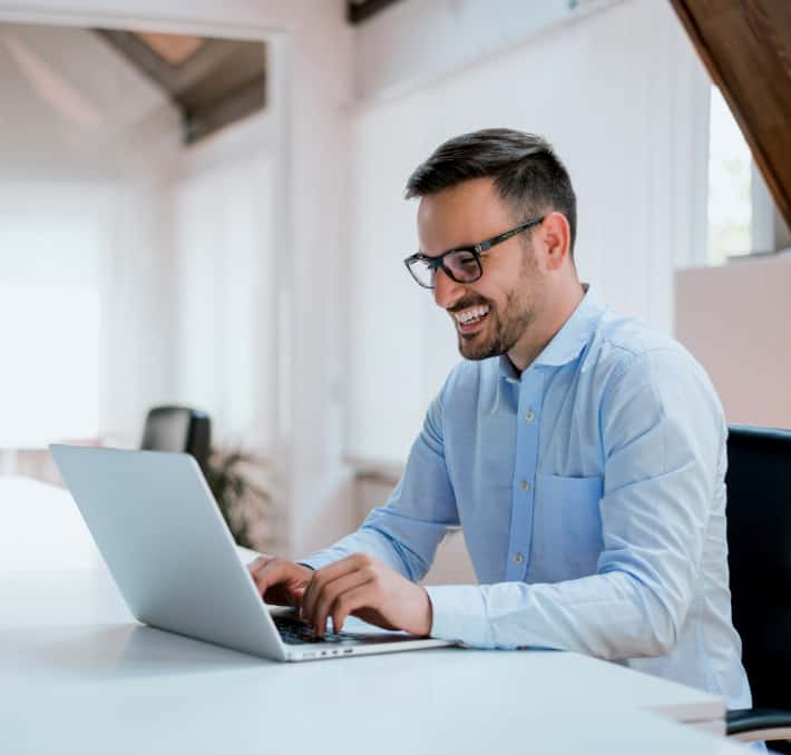 Man in a light blue shirt, wearing glasses, smiles while typing on a laptop at a white desk. The office setting buzzes with B2B e-commerce activity, reflecting the digital ages dynamic business landscape.