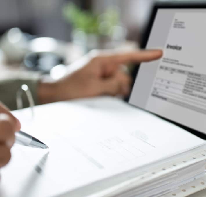 A person points at a digital invoice on a laptop, highlighting the TCO of biomedical devices, while holding a pen over a stack of papers.