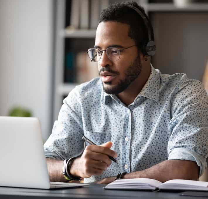 A man with glasses and headphones sits at a desk, using a laptop and holding a pen. An open notebook lies before him as he embarks on the beginning of the Idea-to-Cash journey, ready to transform thoughts into tangible outcomes.