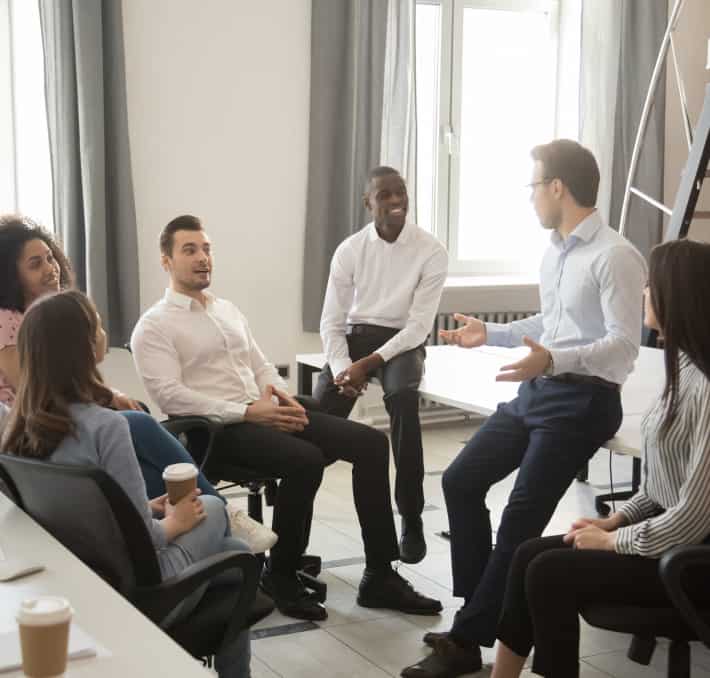 In an office setting, a group sits in a circle, engaged in conversation about emerging technologies. One person gestures animatedly while discussing the potential of digital twins for fluid simulations.