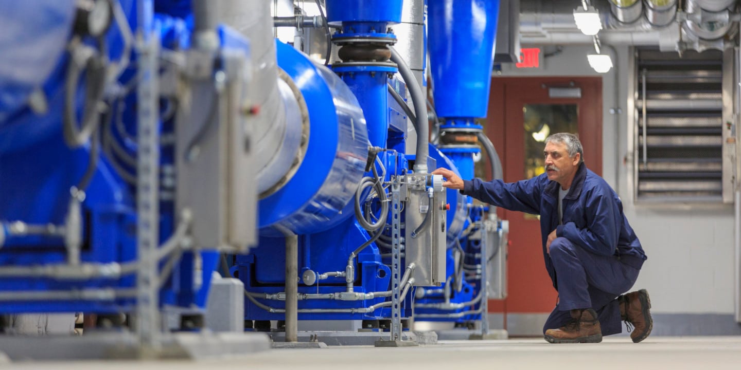 A worker in a blue uniform crouches beside large blue industrial machinery, inspecting a component in a spacious facility. Overhead pipes and metallic structures are visible. The setting is well-lit and organized.