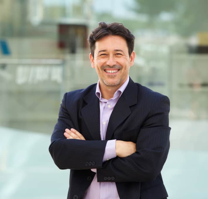 A man in a suit smiles confidently, arms crossed, embodying the perfect synergy of professionalism and innovation against a blurred office backdrop, hinting at the seamless integration of digital twin technology.