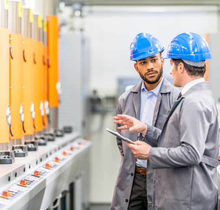 Two men in blue helmets and gray coats discuss machinery in an industrial setting, seamlessly integrating PLM into their conversation. One holds a tablet, demonstrating effective product management techniques, while the other attentively listens.