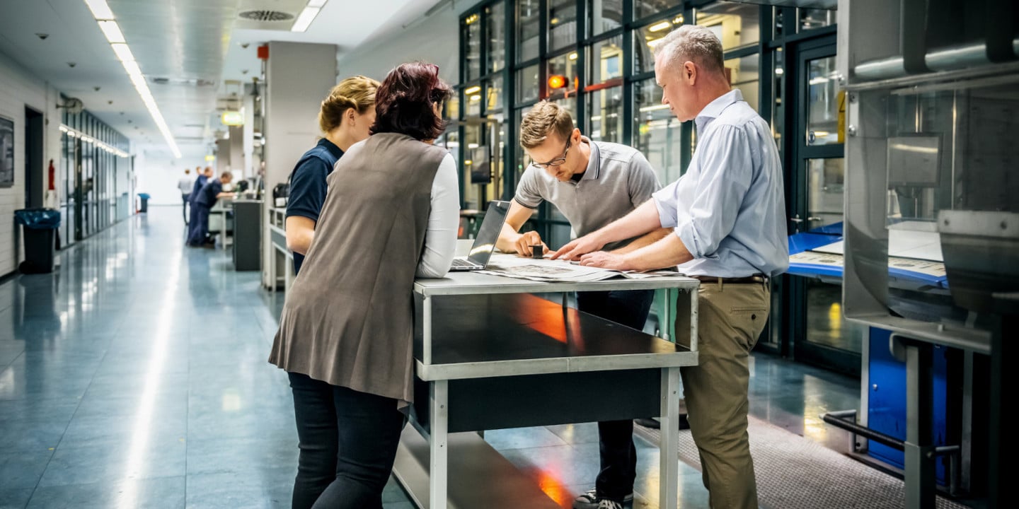 Four people stand around a table in a modern, well-lit office hallway with glass-panelled walls, their focus sharp as they discuss the path to CPQ success while examining various documents.