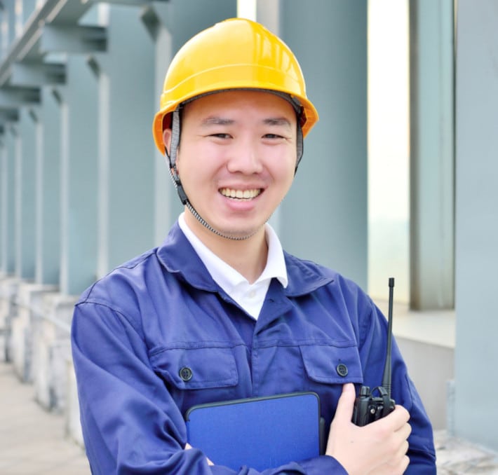 A smiling person in a yellow hard hat and blue work uniform holds a walkie-talkie and tablet, preparing for fluid flow calculations near a building with large windows.