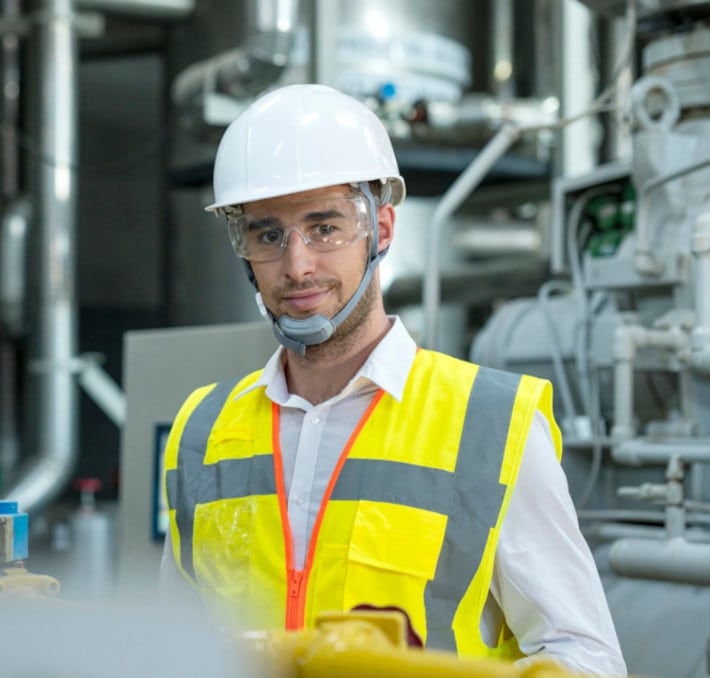 A worker in a bright yellow safety vest and white helmet stands in an industrial setting, expertly overseeing the fluid flow among the intricate machinery in the background.