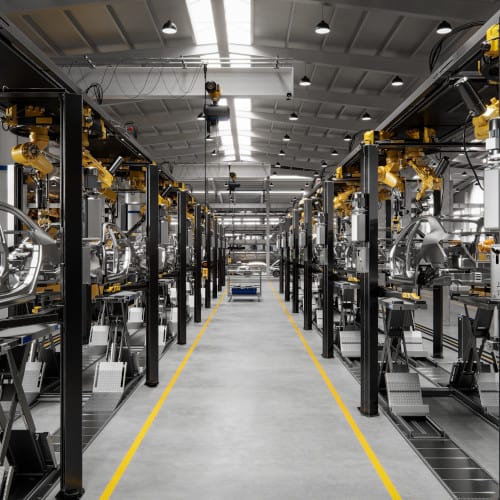 Interior of a modern factory with machinery and equipment aligned along a central aisle, under a high ceiling with skylights.