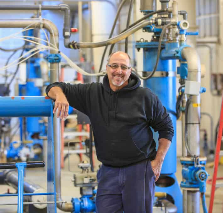 A person wearing a black hoodie stands smiling in an industrial setting, framed by blue machinery and pipes, reminiscent of a water flowing in a gravity-fed system.