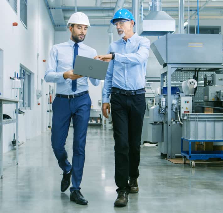 Two men in hard hats and business attire walk through a manufacturing facility, discussing fluid modeling software on a laptop.