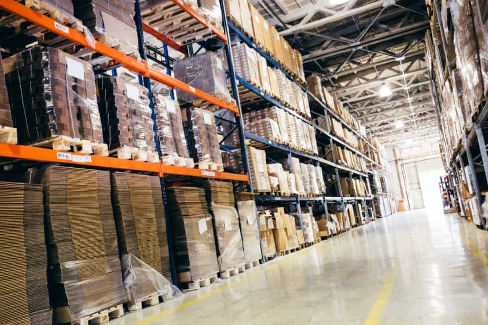 Warehouse interior at eRep Industries showcases rows of metal shelves brimming with diverse boxes and packages. The empty aisle stands ready for activity, while above, exposed beams and lighting illuminate the space we diligently serve.