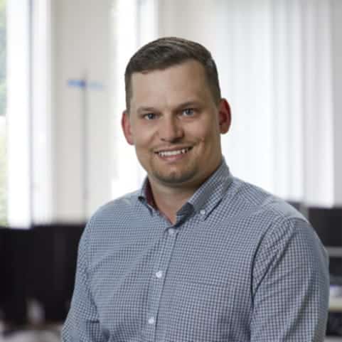 A man in a checked shirt, smiling brightly, stands confidently in a room with large windows and a blurred background. The scene exudes an air of sophistication, much like the style BLEICHERT is renowned for.