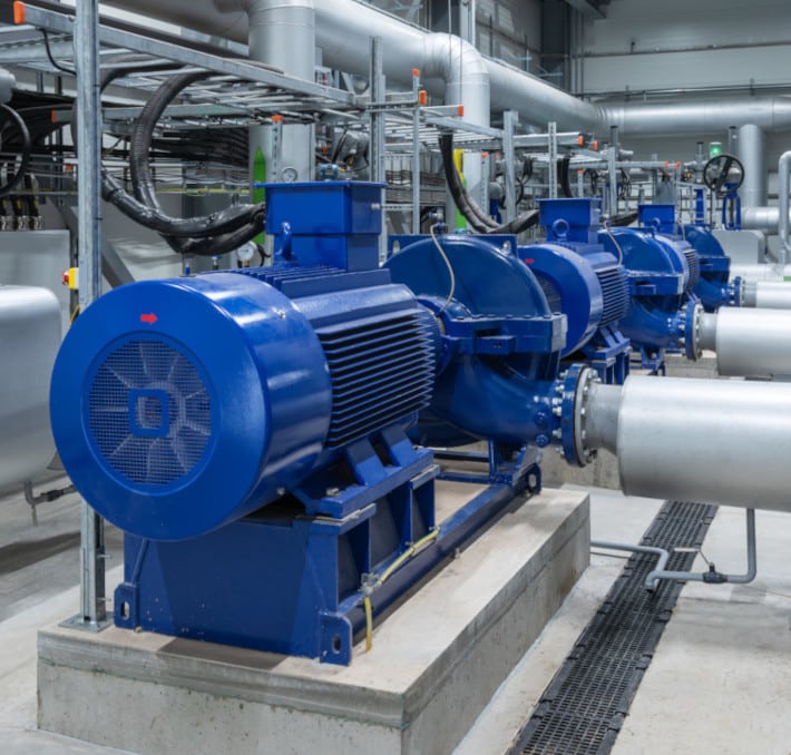 A row of blue industrial pumps and motors, including a robust Net Positive Suction Head pump, operates in a facility, seamlessly connected to large metal pipes.