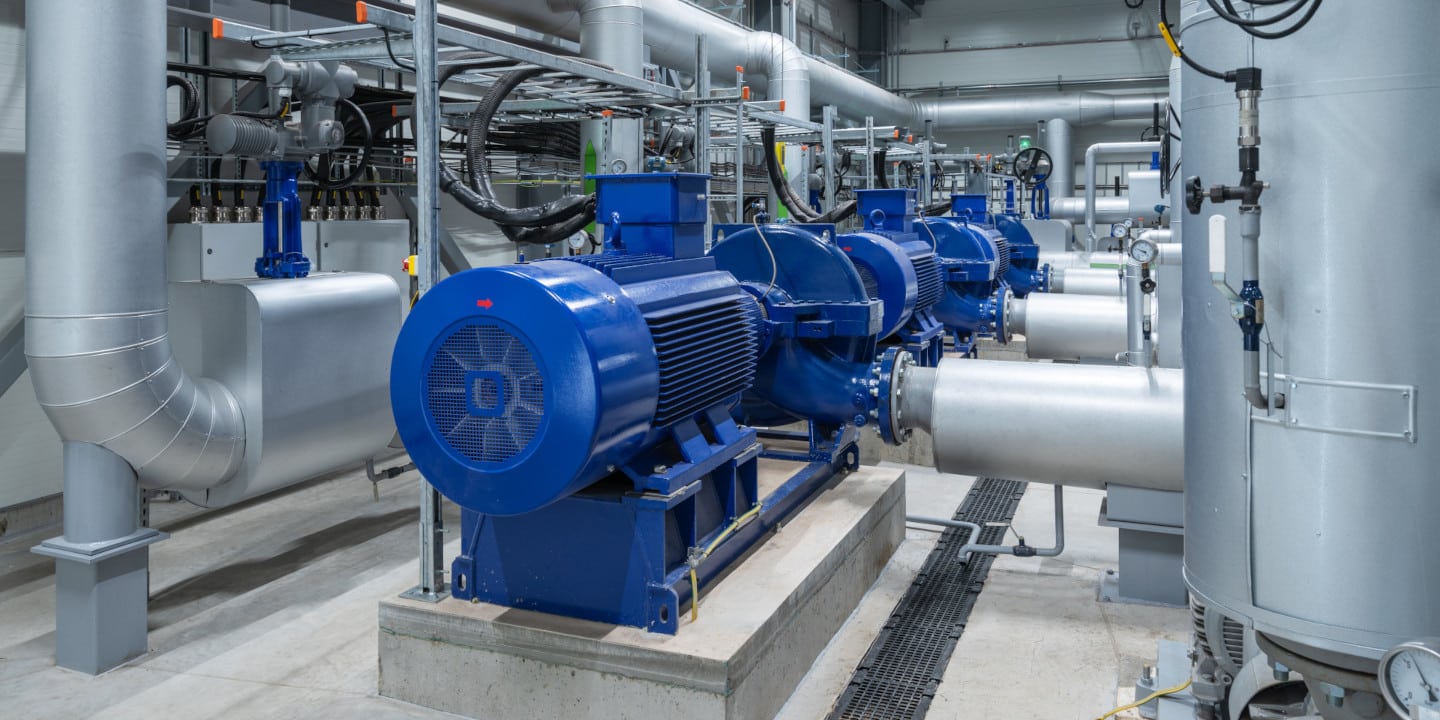 In an industrial setting, large blue machinery and interconnected pipes dominate the concrete floor, while various cables and valves weave through. A Net Positive Suction Head Pump hums efficiently, ensuring optimal fluid dynamics amidst the organized chaos.
