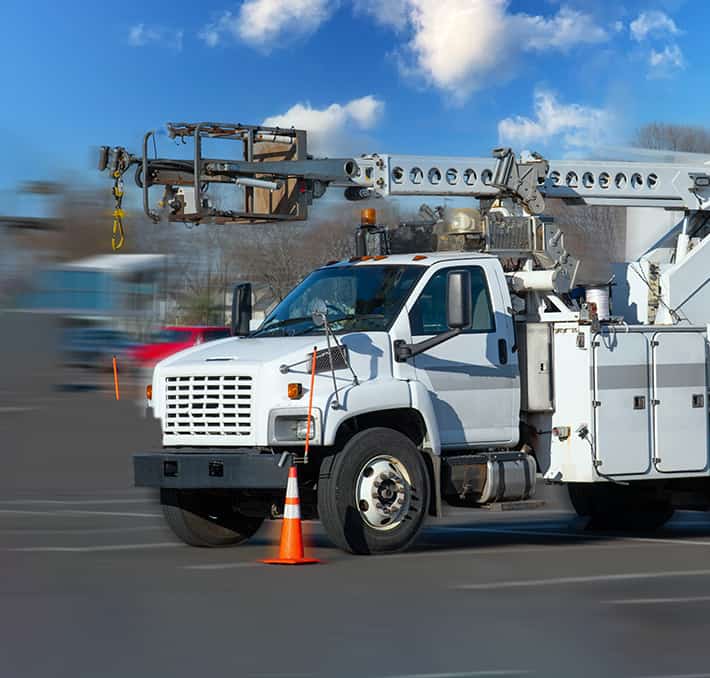 A white utility truck, crafted with excellence in specialty vehicle manufacturing, features an extended boom lift and is parked beside an orange traffic cone on a paved surface under a blue sky dotted with clouds.