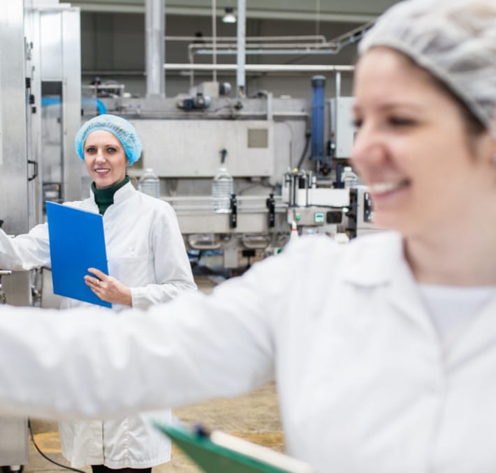 Two workers in white lab coats and hairnets hold clipboards in an industrial food processing facility, ensuring strict Food & Beverage Compliance.