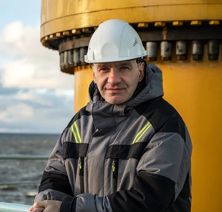 A person wearing a white helmet and gray jacket stands outdoors near a large, yellow industrial structure, likely part of an extensive fluid handling system.