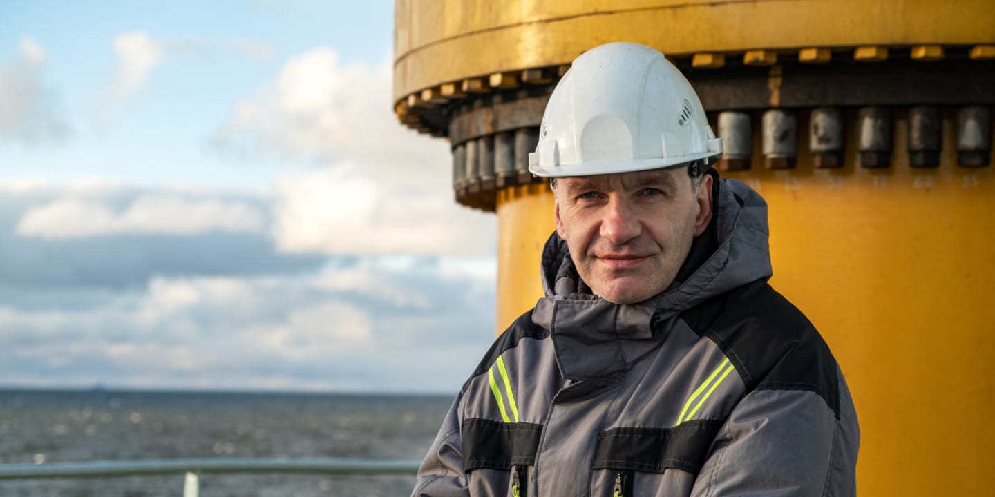 A person wearing a white hard hat and a gray jacket stands in front of a large yellow fluid handling system by the sea.
