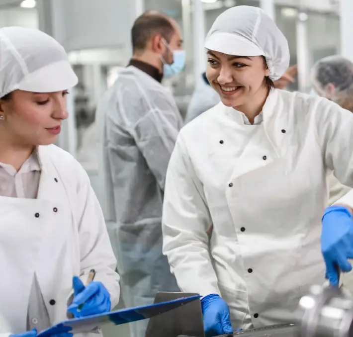 Two workers in white uniforms and hairnets, wearing blue gloves, discuss food and beverage industry challenges in a lab or processing facility. Others in similar attire are visible in the background, focused on maintaining high standards amidst these evolving issues.
