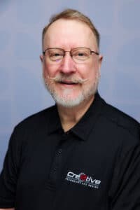 Ein Mann mit Brille und Schnurrbart, der ein schwarzes Polohemd mit einem Creative Technology and Design-Logo trägt, steht vor einem hellblauen Hintergrund und verkörpert den Wettbewerbsvorteil durch PLM-Know-how.