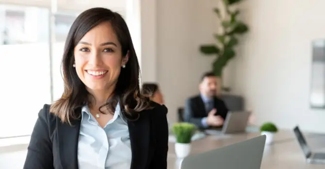 In a modern office setting, a woman smiles at the camera while her colleagues are immersed in discussions about AQ CPQ for Mac in the background.