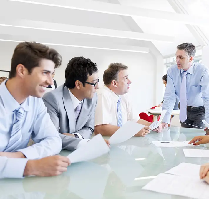 Five people in business attire are gathered around a glass table for a meeting in a bright office setting, discussing the latest updates on TENADO Service with papers in hand.
