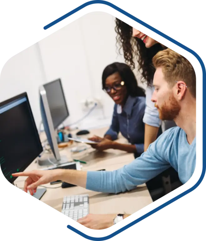 In a bustling office, three people gather around computers. A man with a beard points at the screen, leading the conversation during a TENADO Training session, while a woman with glasses and another woman engage intently.