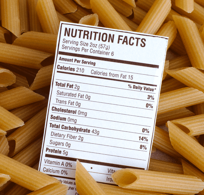 A nutrition facts label for pasta is placed on top of uncooked penne pasta, displaying nutritional information per serving.