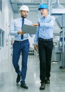 Two men in safety helmets walk through an industrial facility, one holding a laptop open to monitor PLM metrics for product development.