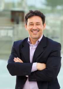 A man in a suit smiles confidently, arms crossed, embodying the perfect synergy of professionalism and innovation against a blurred office backdrop, hinting at the seamless integration of digital twin technology.