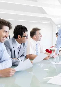Five people in business attire are gathered around a glass table for a meeting in a bright office setting, discussing the latest updates on TENADO Service with papers in hand.