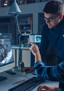Two people at a desk engage in a focused discussion about a 3D mechanical design displayed on a computer monitor. In an office setting, they work collaboratively, building the digital thread that seamlessly integrates ideas into the design process.